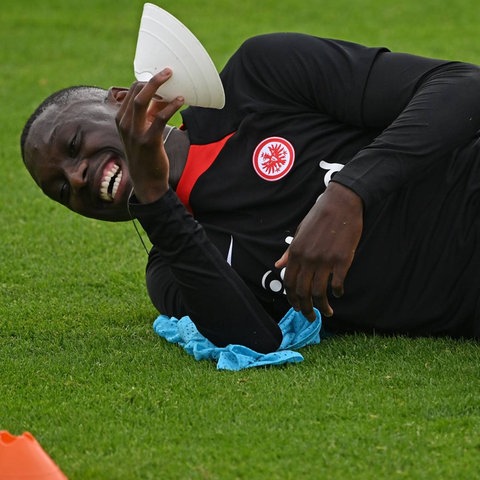 Eintracht-Spieler Kolo Muani liegt auf dem Rasen und lacht. Rechts am Bildrand ist ein Foto von hr-Sportreporter Philipp Hofmeister montiert. Darauf trägt er ein blaues Hemd und hält ein blaues ARD-Mikrofon in der Hand.